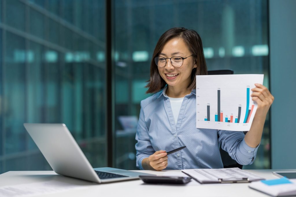 Asian businesswoman analyzing financial growth with a laptop and charts in modern office setting, displaying positive results during business presentation