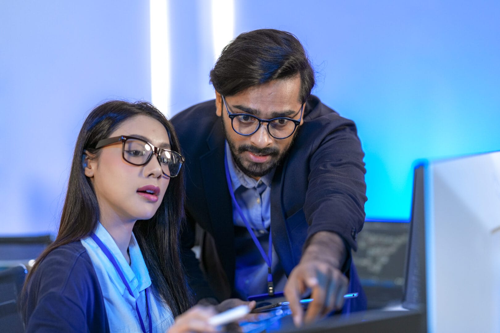 Diverse developers collaborate, using a desktop for coding and a tablet for input. A man and woman review UI/UX design and code in a futuristic, blue-lit tech office.