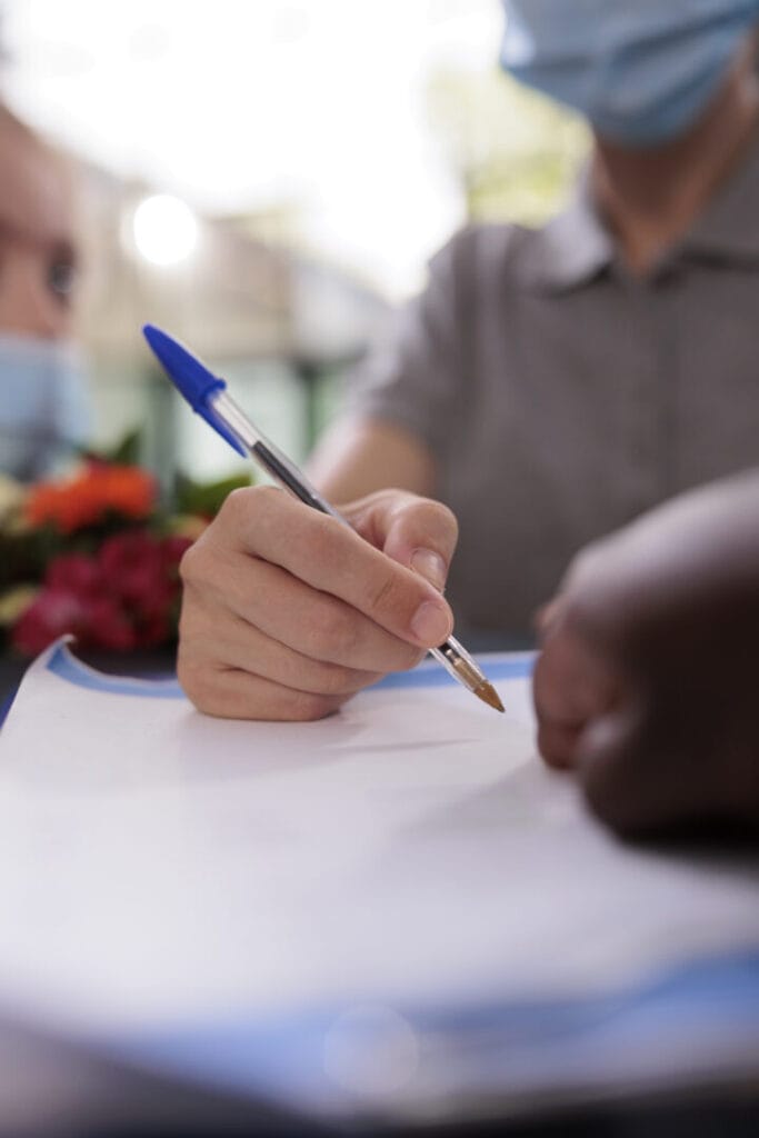 Close up of mother hand signing insurance documents before start consultation with physician, family standing in hospital waiting area. People wearing protection face mask against coronavirus